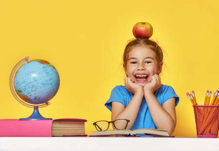Back to school and happy time! Cute industrious child is sitting at a desk indoors. Kid is learning in class on background of bright yellow wall. Girl reading the book.の写真素材