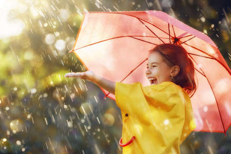 Happy funny child with red umbrella under the autumn shower. Girl is wearing yellow raincoat and enjoying rainfall. Kid playing on the nature outdoors. Family walk in the park.の写真素材