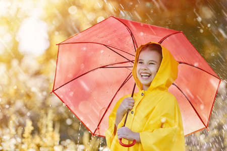 Happy funny child with red umbrella under the autumn shower. Girl is wearing yellow raincoat and enjoying rainfall. Kid playing on the nature outdoors. Family walk in the park.の写真素材