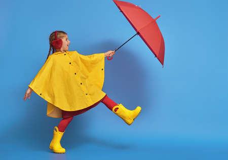 Happy funny child with red umbrella posing on blue wall background. Girl is wearing yellow raincoat and rubber boots.の写真素材