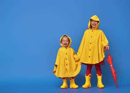 Two happy funny children with red umbrella posing on blue wall background. Girls is wearing yellow raincoat and rubber boots.の写真素材