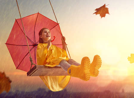 Happy funny child with red umbrella under the autumn shower. Girl is wearing yellow raincoat, rubber boots and enjoying rainfall. Kid playing on the nature outdoors. Family walk in the park.の写真素材