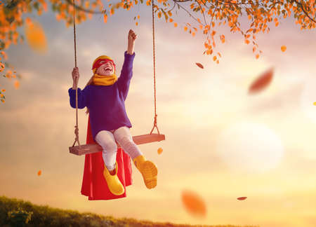 Happy child on swing in sunset fall. Little kid playing in the autumn on the nature walk. Girl in superhero costume.の写真素材