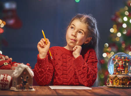 Merry Christmas and Happy Holidays! Cute little child girl is writing the letter to Santa Claus near...
