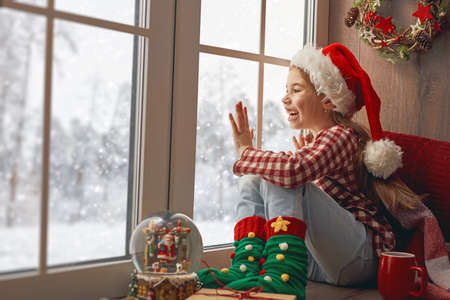 Merry Christmas and happy holidays! Cute little girl sitting by the window with a cup of hot drink and looking at the winter forest. Room decorated. Kid enjoys the snowfall.の写真素材