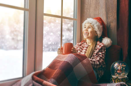 Merry Christmas and happy holidays! Cute little girl sitting by the window with a cup of hot drink and looking at the winter forest. Room decorated. Kid enjoys the snowfall.の写真素材