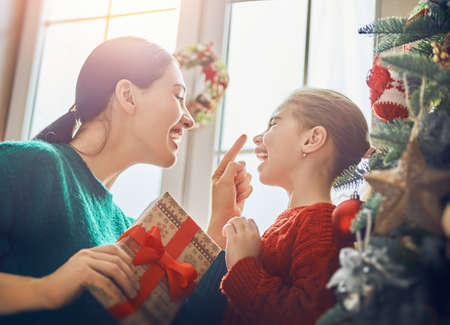 Merry Christmas and Happy Holidays! Cheerful mom and her cute daughter girl exchanging gifts. Parent and little child having fun near tree indoors. Morning Xmas. Portrait family close up.の写真素材
