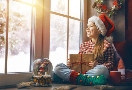 Merry Christmas and happy holidays! Cute little girl sitting by the window with a present gift box and looking at the winter forest. Room decorated. Kid enjoys the snowfall.の写真素材