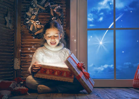 Merry Christmas and Happy Holiday! Cute little child girl with magical present gift box sitting by the window.の写真素材