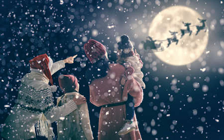 Merry Christmas and happy holidays! Cute little children with mom and dad. Santa Claus flying in his sleigh against moon sky. Family enjoying the holiday on dark background.の写真素材