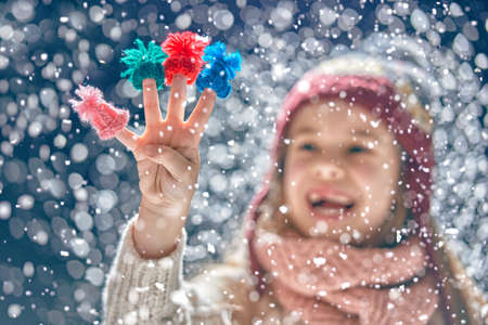 Winter portrait of happy little girl wearing knitted hat, scarf and sweater. Child having small hats on her fingers on dark snowy background. Family fashion concept.の写真素材