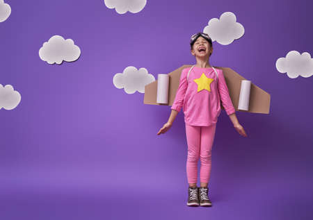 Little child girl in an astronaut costume is playing and dreaming of becoming a spaceman. Portrait of funny kid on a background of ultraviolet wall with white clouds.の写真素材