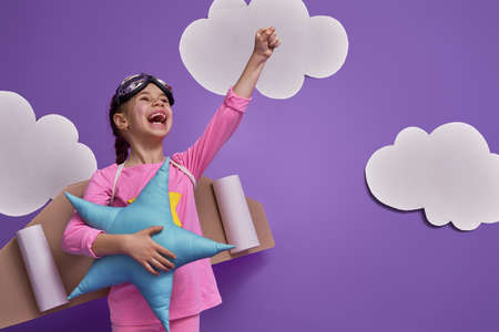 Little child girl in an astronaut costume is playing and dreaming of becoming a spaceman. Portrait of funny kid on a background of ultraviolet wall with white clouds.の写真素材