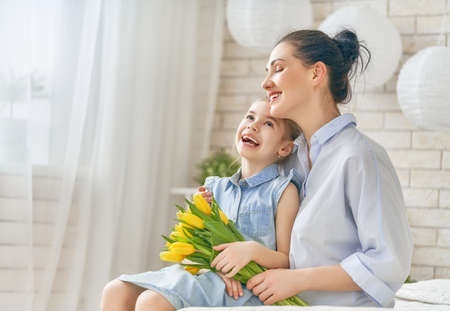 Happy women's day! Child daughter is congratulating mom and giving her flowers tulips. Mum and girl smiling and hugging. Family holiday and togetherness.の写真素材