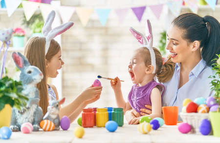 Happy holiday! A mother and her daughters are painting eggs. Family preparing for Easter. Cute little children girls are wearing bunny ears.の写真素材