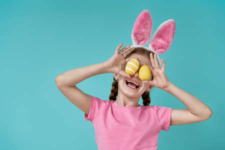 Cute little child wearing bunny ears on Easter day. Girl holding basket with painted eggs on light blue background.の写真素材