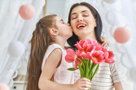 Happy women's day! Child daughter is congratulating mom and giving her flowers tulips. Mum and girl smiling and hugging. Family holiday and togetherness.の写真素材