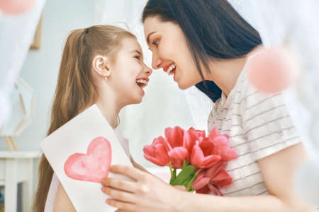 Happy women's day! Child daughter is congratulating mom and giving her flowers tulips. Mum and girl smiling and hugging. Family holiday and togetherness.の写真素材