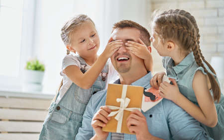 Happy father's day! Children daughters congratulating dad and giving him postcard and gift box. Daddy and girls smiling and hugging. Family holiday and togetherness.の写真素材