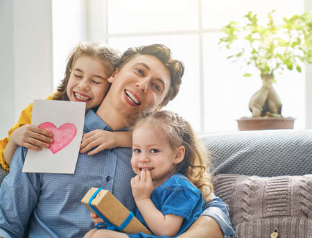 Happy father's day! Children daughters congratulating dad and giving him postcard and gift box. Daddy and girls smiling and hugging. Family holiday and togetherness.の写真素材