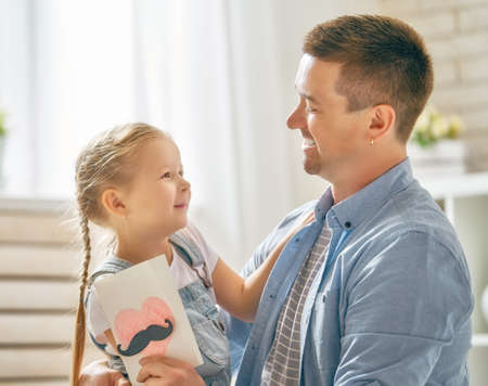 Happy father's day! Child daughter congratulating dad and giving him postcard. Daddy and girl smiling and hugging. Family holiday and togetherness.の写真素材