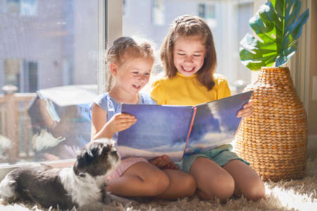 Cute children girls are reading a book sitting on floor in room at home.の写真素材