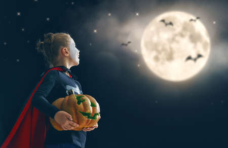 Happy Halloween! Cute little skeleton with pumpkin. Beautiful young child girl in costume looking at full moon.の写真素材