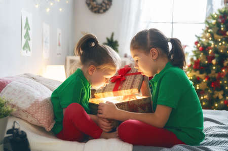 Merry Christmas and Happy Holidays! Cheerful cute childrens girls opening gifts on the bed. Kids wearing pajamas having fun near tree in the morning. Loving family with presents in room.の写真素材