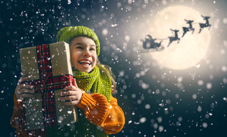Merry Christmas! Cute little child with xmas present. Santa Claus flying in his sleigh against moon sky. Happy kid enjoy the holiday. Portrait of girl with gifts on dark background.の写真素材