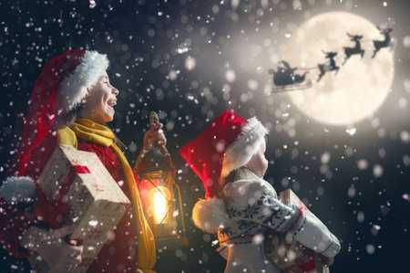 Merry Christmas and happy holidays! Cute little children with xmas presents. Santa Claus flying in his sleigh against moon sky. Kids enjoying the holiday with gifts on dark background.の写真素材