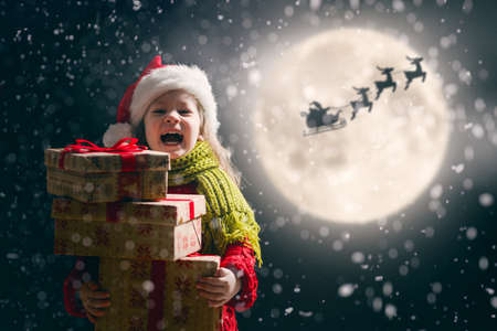 Merry Christmas! Cute little child with xmas present. Santa Claus flying in his sleigh against moon sky. Happy kid enjoy the holiday. Portrait of girl with gifts on dark background.の写真素材