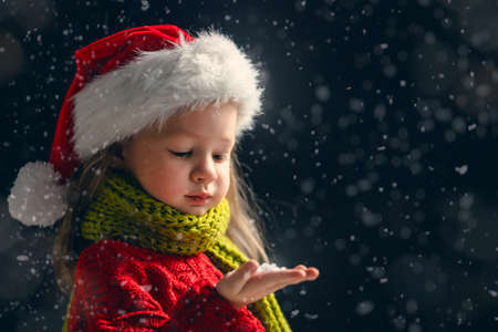 Merry Christmas and happy holidays! Little girl blowing on snow. Child enjoying the game outdoors. Portrait kid on dark background.の写真素材
