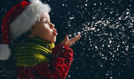 Merry Christmas and happy holidays! Little girl blowing on snow. Child enjoying the game outdoors. Portrait kid on dark background.の写真素材