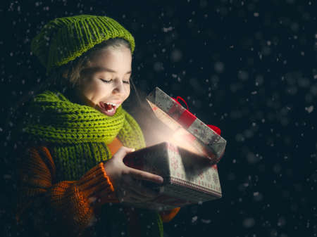 Happy child girl playing on a snowy winter walk. Little girl enjoys the game. Child girl playing outdoors in snow. Outdoor fun for winter vacation. Portrait kid with gift box on dark background.の写真素材