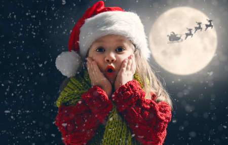 Cute little child on Christmas. Santa Claus flying in his sleigh against moon sky. Happy kid enjoy the holiday. Portrait of girl with gifts on dark background.の写真素材