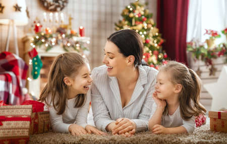 Merry Christmas and Happy Holidays! Cheerful mom and her cute daughters girls exchanging gifts. Parent and little children having fun near tree indoors. Loving family with presents in room.の写真素材