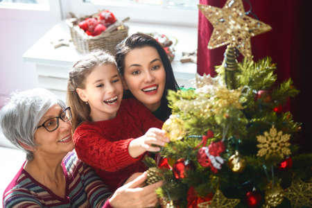 Merry Christmas and Happy Holidays! Grandma, mom and daughter decorate the tree indoors. The morning...