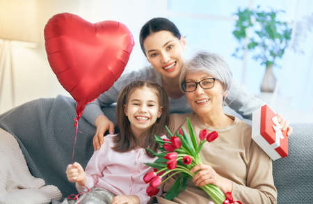 Happy women's day! Child daughter is congratulating mom and granny giving them flowers tulips. Grandma, mum and girl smiling and hugging. Family holiday and togetherness.の写真素材