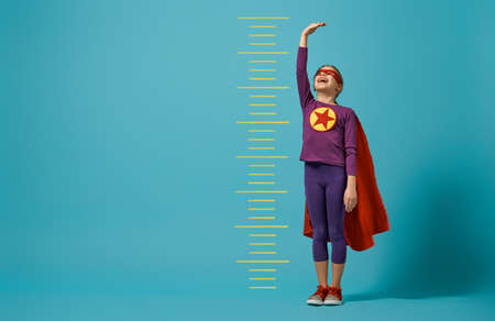 Little child playing superhero. Kid measures the growth on the background of bright blue wall. Girl power concept. Red, purple and  turquoise colors.の写真素材