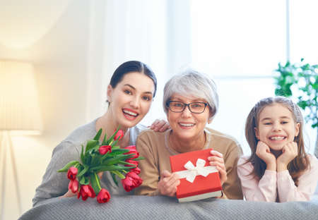 Happy women's day! Child daughter is congratulating mom and granny giving them flowers tulips. Grandma, mum and girl smiling and hugging. Family holiday and togetherness.の写真素材