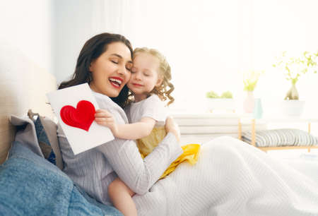 Happy mother's day! Child daughter is congratulating mom and giving her postcard and gift. Mum and girl smiling and hugging. Family holiday and togetherness.の写真素材