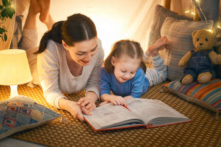 Family bedtime. Mom and child daughter are reading a book in tent. Pretty young mother and lovely girl having fun in children room.の写真素材