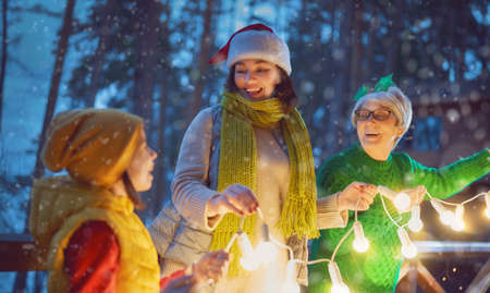Merry Christmas and Happy Holidays! Cheerful mom, granny and cute girl decorating home. Parents and little child having fun outdoors. Loving family with garlands outside.の写真素材