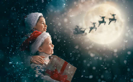 Merry Christmas and happy holidays! Cute little children with xmas presents. Santa Claus flying in his sleigh against moon sky. Kids enjoying the holiday with gifts on dark background.の写真素材