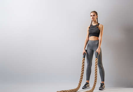 Athletic young woman doing crossfit exercises with a rope on grey background.