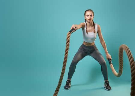 Athletic young woman doing crossfit exercises with a rope on teal background.