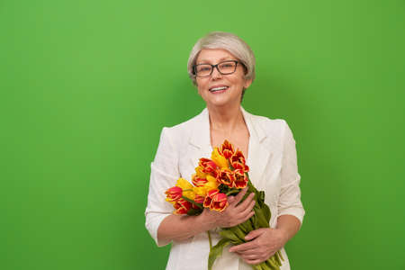 Beautiful senior woman with yellow flowers tulips in hands on color green background.の写真素材