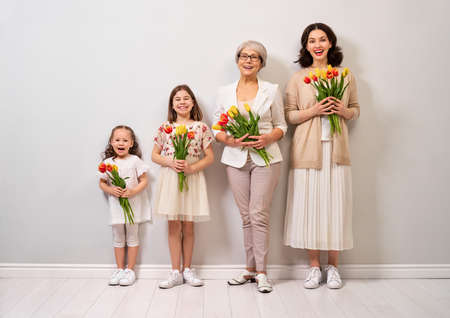 Happy women's day! Children daughters are congratulating mom and grandma giving them flowers tulips.Granny, mum and girls smiling on light grey background. Family holiday and togetherness.の写真素材