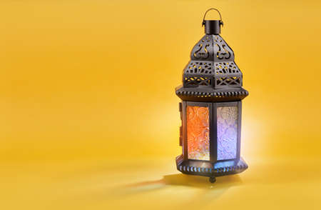Ornamental Arabic lantern with burning candle glowing on yellow background. Festive greeting card,...