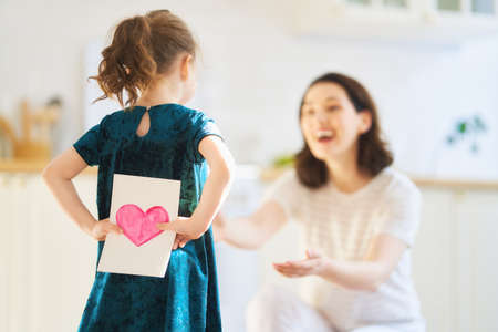 Happy mother's day! Child daughter is congratulating mom and giving her postcard. Mum and girl smiling and hugging. Family holiday and togetherness.の写真素材